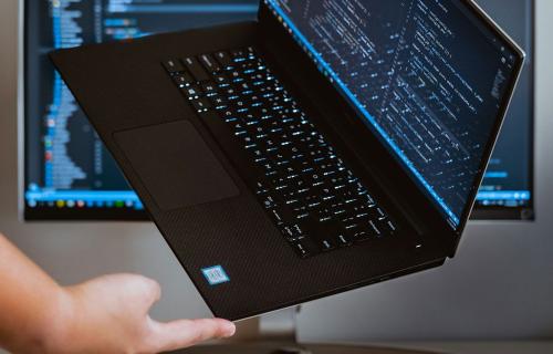 A hand holds open a laptop displaying lines of code, with two monitors showing code and data visualizations in the background.